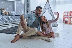 selfie engraçada. jovem pai e sua filha tomando selfie enquanto está sentado no chão no quarto com a tenda ao fundo foto