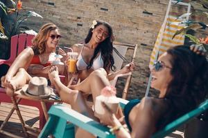 mulheres jovens atraentes em biquíni bebendo coquetéis e sorrindo enquanto relaxam ao ar livre foto