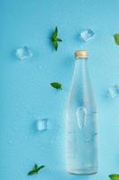 garrafa com uma bebida gelada, cubos de gelo, gotas e folhas de hortelã em um fundo azul. foto