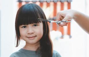 mãos femininas para cortar o cabelo da frente ou franja para uma linda garota asiática em casa. as mães ficam felizes em cortar o cabelo de seus filhos. conceito de cuidados com o cabelo foto