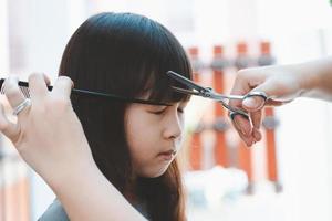 mão feminina corta cabelo para menina asiática com tesoura e pente. conceito de cuidados com o cabelo foto