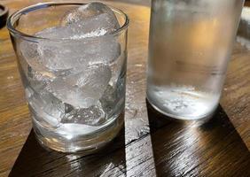 foco seletivo, um copo transparente cheio de gelo e garrafa de vidro transparente contém água mineral fria se senta na mesa de madeira foto