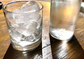 foco seletivo, um copo transparente cheio de gelo e garrafa de vidro transparente contém água mineral fria se senta na mesa de madeira foto