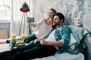 muito divertido lindo casal jovem tomando selfie e fazendo caretas enquanto passa o tempo na cama em casa foto