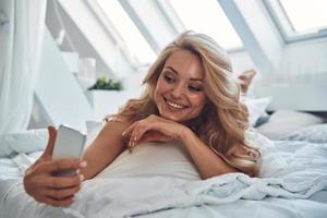 bela jovem sorridente usando telefone inteligente enquanto estava deitado na cama em casa foto