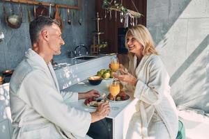 lindo casal maduro em roupões de banho tomando café da manhã juntos enquanto passa o tempo na cozinha doméstica