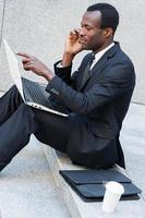 ocupado trabalhando ao ar livre. Vista lateral do jovem africano confiante em trajes formais falando no celular e trabalhando no laptop enquanto está sentado ao ar livre foto