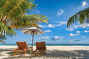 praia de férias incrível. cadeiras na praia de areia perto do mar. turismo de férias românticas de verão. bela paisagem de ilha tropical. cenário tranquilo da costa, relaxe o horizonte à beira-mar de areia, folhas de palmeira foto