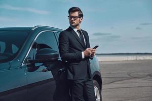 negócio é a vida dele. bonito jovem empresário usando telefone inteligente em pé perto de seu carro ao ar livre foto