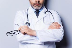 confiança e experiência. close-up do médico de cabelos grisalhos maduros, mantendo os braços cruzados e segurando óculos em pé contra um fundo cinza foto