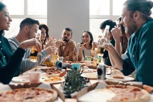 ótima festa. grupo de jovens em roupas casuais comendo pizza e sorrindo enquanto faz um jantar dentro de casa foto