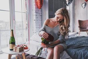 suas flores favoritas. mulher jovem e atraente segurando um buquê de rosas vermelhas e sorrindo enquanto está sentado na cama em casa foto