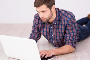 homem navegando na net. jovem bonito trabalhando no laptop enquanto estava deitado no chão de madeira foto