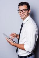examinando seu novo tablet digital. Vista lateral do jovem bonito de camisa e gravata trabalhando em tablet digital e sorrindo em pé contra um fundo cinza foto