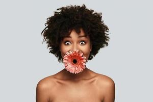 o que surpreendeu a jovem africana olhando para a câmera e mantendo a flor na boca em pé contra um fundo cinza foto