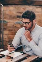 verificando-o duas vezes. bonito jovem pensativo segurando o telefone inteligente e olhando para ele enquanto está sentado em seu local de trabalho foto