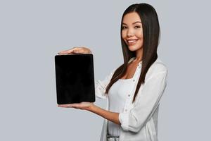 você deveria ver isso. atraente jovem mulher asiática apontando espaço de cópia em seu tablet digital e sorrindo em pé contra um fundo cinza foto