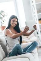 linda e confiante. mulher jovem e atraente segurando um tablet digital e sorrindo enquanto relaxa no sofá em casa foto