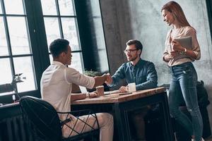 jovens colegas modernos em roupas casuais inteligentes apertando as mãos e sorrindo enquanto trabalhavam no escritório foto