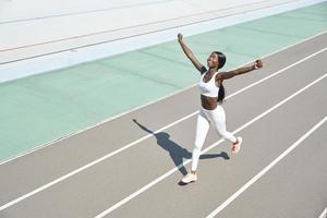 vista superior da jovem africana feliz em roupas esportivas correndo na pista e mantendo os braços levantados foto