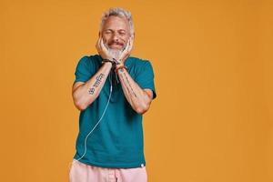 feliz homem sênior ouvindo música e sorrindo em pé contra um fundo laranja foto