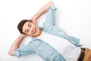 tão feliz e despreocupado. vista superior do jovem alegre segurando as mãos atrás da cabeça e sorrindo enquanto estava deitado no branco foto