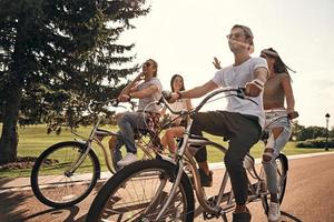 não há necessidade de andar se você tiver rodas. grupo de jovens felizes em roupas casuais sorrindo enquanto andam de bicicleta juntos ao ar livre foto