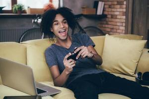 jogos sempre divertidos. alegre jovem africano jogando videogame e mantendo a boca aberta enquanto está sentado no sofá em casa foto