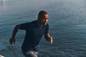 dando todo o seu melhor. jovem africano em roupas esportivas correndo enquanto se exercita perto do rio ao ar livre foto