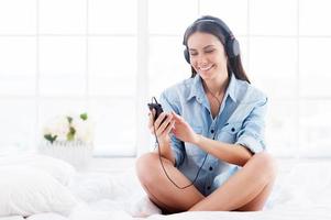 curtindo sua música favorita. mulher jovem e bonita em fones de ouvido ouvindo mp3 player e sorrindo enquanto está sentado na cama foto