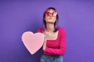 linda mulher segurando coração de papel e mandando um beijo em pé contra um fundo roxo foto