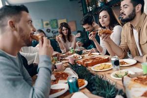 bons momentos com os amigos. grupo de jovens em roupas casuais comendo pizza e sorrindo enquanto faz um jantar dentro de casa foto