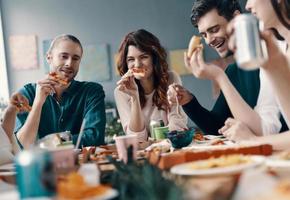 deixar todas as preocupações para trás. grupo de jovens em roupas casuais comendo pizza e sorrindo enquanto faz um jantar dentro de casa foto