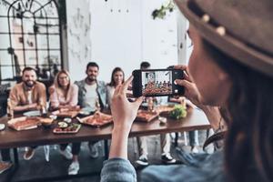 criando memórias felizes. jovem fotografando um grupo de amigos usando telefone inteligente enquanto eles fazem um jantar foto