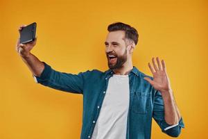 jovem feliz em roupas casuais tomando selfie usando telefone inteligente e sorrindo em pé contra um fundo amarelo foto