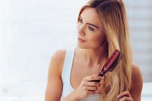 pentear o cabelo para mantê-lo saudável. close-up de mulher jovem e bonita escovando seus longos cabelos e olhando para longe em pé contra a parede de tijolos brancos foto