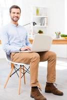 fácil de trabalhar. jovem bonito na camisa trabalhando no laptop na frente de sua mesa e sorrindo para a câmera enquanto está sentado no escritório foto