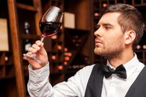 este vinho é simplesmente perfeito. sommelier masculino confiante examinando o copo com vinho em pé perto da prateleira de madeira foto