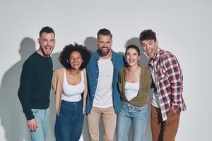 grupo de jovens bonitos em roupas casuais, unindo-se e sorrindo em pé contra um fundo cinza foto