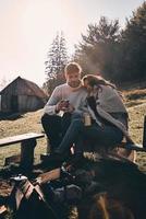 aproveitando cada minuto juntos. lindo casal jovem tomando café da manhã enquanto está sentado junto à fogueira nas montanhas foto