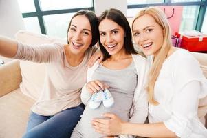 foco na felicidade. vista superior da jovem grávida feliz segurando botinhas de bebê no abdômen e sorrindo enquanto dois amigos sentados perto dela e fazendo selfie foto