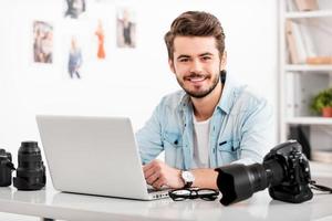 fotógrafo jovem e criativo. jovem sorridente trabalhando no laptop e olhando para a câmera enquanto está sentado em seu local de trabalho foto