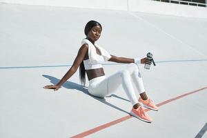 bela jovem africana em roupas esportivas, segurando a garrafa com água enquanto está sentado na pista de esportes ao ar livre foto