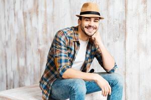 casualmente bonito. jovem bonito usando chapéu e olhando para a câmera enquanto está sentado contra a parede de madeira foto