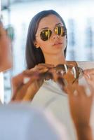 fazendo sua escolha final. mulher jovem e bonita escolhendo óculos de sol para comprar em frente ao espelho na loja de óptica foto