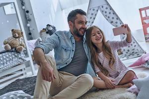 tempo selfie pai alegre e sua filha tomando selfie enquanto está sentado no chão no quarto com a tenda ao fundo foto