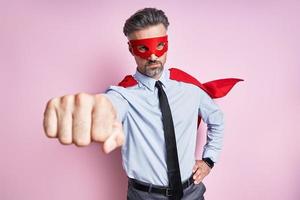 homem de camisa e gravata vestindo capa de super-herói e mantendo o braço estendido contra o fundo rosa foto