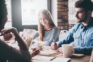 ótimas notícias jovem alegre olhando para a câmera com sorriso enquanto está sentado à mesa do escritório na reunião de negócios com seus colegas de trabalho foto