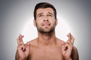 implorando por... retrato de jovem sem camisa, mantendo os dedos cruzados e olhando para cima em pé contra um fundo cinza foto