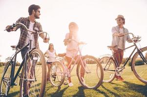 passar bons momentos com os melhores amigos. jovens alegres empurrando suas bicicletas enquanto caminhavam pela grama verde ao ar livre foto